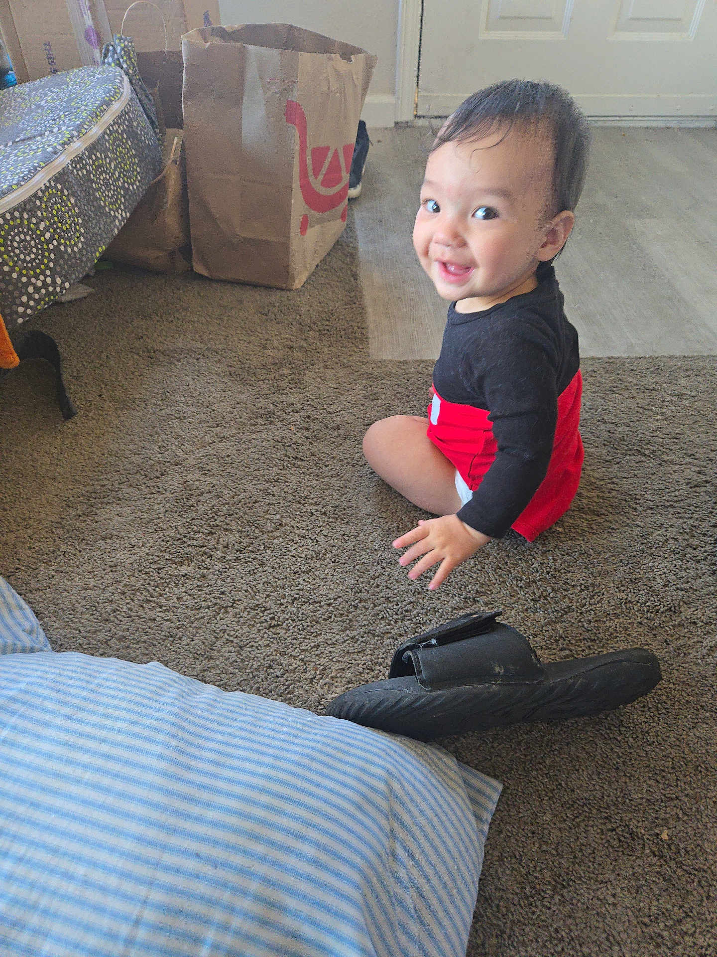 Keanan is registered to the contest to win money with this photo: toddler, child, smiling, carpet, slipper, pillow, paper_bag, door, floor, indoor, clothing, person, home, happy, sitting, black_shirt, red_shorts, head, face, light
