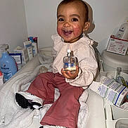 Layanah a rejoint le concours — aidez-le/la à gagner de superbes lots ! toddler, child, smiling, sitting, white_blanket, bottle, baby_products, shoes, pink_pants, long_sleeve_shirt, indoor, face, happy, skin_condition, cute, person, baby_care, table, cozy, joyful