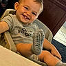 baby, child, smiling, portrait, high_chair, indoor, gray_shirt, socks, barefoot, happy, teeth, blond_hair, seated, close_up, candid, home, furniture, warm_lighting, playful, person