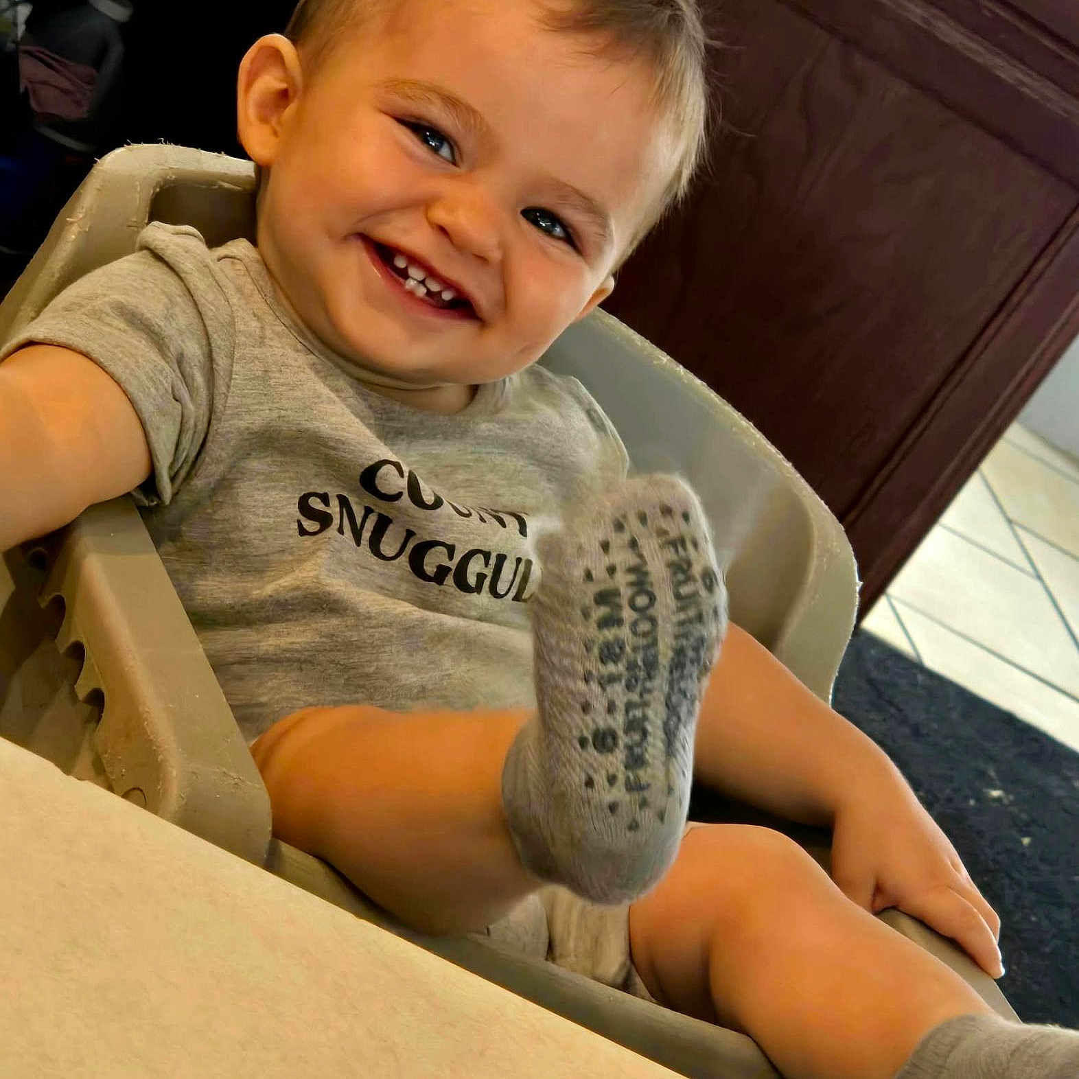 Maverick joined the competition — help win amazing prizes! baby, child, smiling, portrait, high_chair, indoor, gray_shirt, socks, barefoot, happy, teeth, blond_hair, seated, close_up, candid, home, furniture, warm_lighting, playful, person