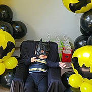 Isaac is registered to the contest to win money with this photo: baby, batman_costume, child, balloons, yellow, black, party, gift_bags, mask, cape, costume, indoor, carpet, chair, celebration, superhero, toddler, cute, fun, portrait