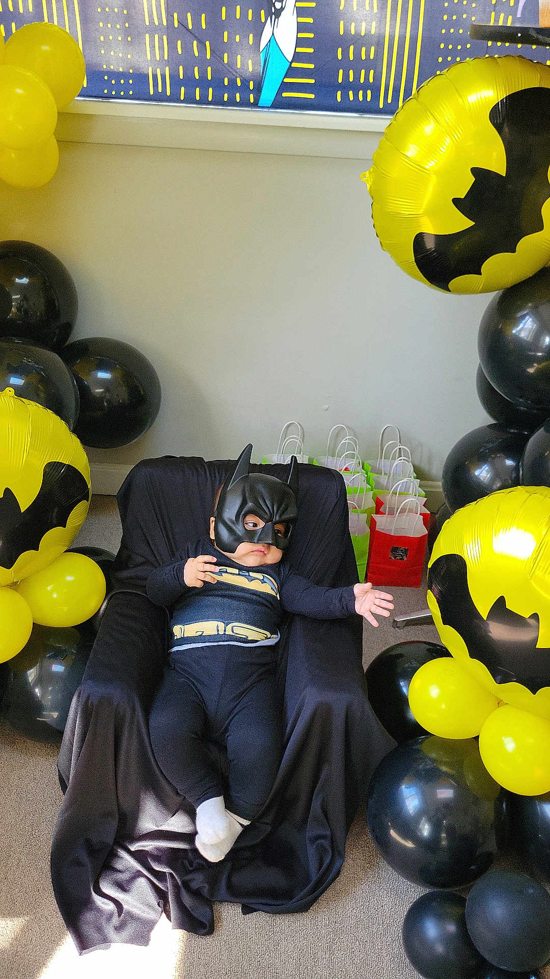 Isaac is registered to the contest to win money with this photo: baby, batman_costume, child, balloons, yellow, black, party, gift_bags, mask, cape, costume, indoor, carpet, chair, celebration, superhero, toddler, cute, fun, portrait