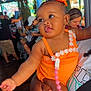 accessory, baby, bow, celebration, child, close_up, clothing, colorful_lights, crowd, cute, family, flower_decorations, hand, happy_expression, indoor, orange_dress, pacifier_clip, people, person_holding, portrait
