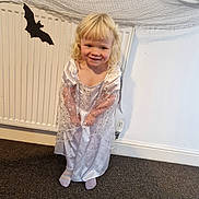 Luna is registered to the contest to win money with this photo: child, blonde_hair, dress, princess_costume, socks, carpet, radiator, bat_decoration, spider_web, indoor, smiling, standing, wall, heater_valve, curly_hair, cute, happy, costume, holiday, playful