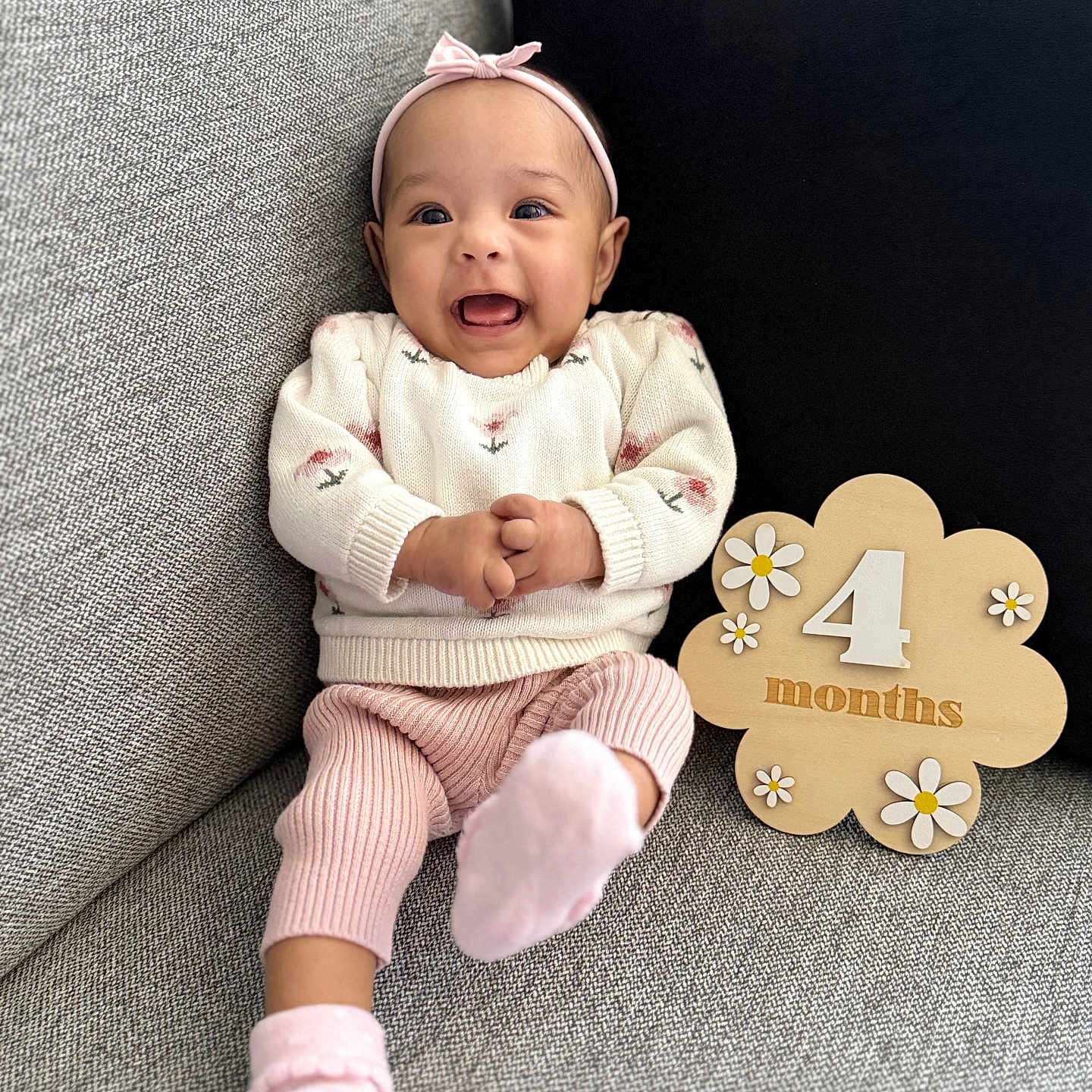 Ylona Margaret joined the competition — help win amazing prizes! 4_months, baby, black_cushion, child, couch, cozy, cute, flower_pattern, gray_couch, happy, headband, indoor, infant, pink_pants, pink_socks, portrait, sitting, smiling, white_sweater, wooden_sign
