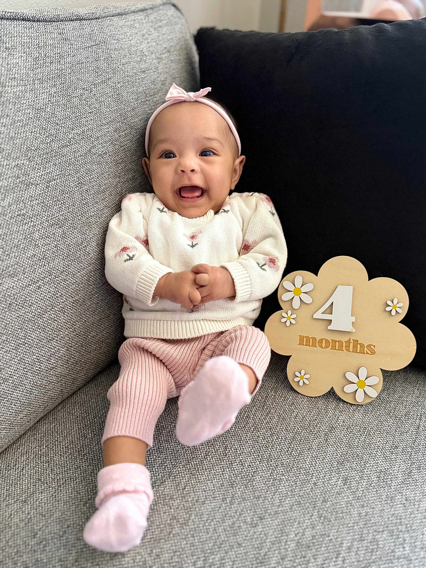 Ylona Margaret joined the competition — help win amazing prizes! baby, infant, smiling, headband, pink_socks, pink_pants, white_sweater, flower_pattern, wooden_sign, 4_months, couch, gray_couch, black_cushion, happy, portrait, indoor, cute, child, sitting, cozy