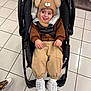 toddler, child, stroller, baby, hat, bear_hat, brown_clothing, pants, white_sneakers, indoor, floor_tiles, smiling, tongue_out, cute, seatbelt, shopping, clothing_rack, footwear, playful, person
