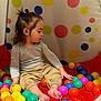 toddler, child, ball_pit, colorful_balls, indoor, playtime, toy, cute, casual_clothing, feet, sitting, hair, gray_shirt, beige_pants, plastic_balls, play_area, young_child, fun, portrait, curious