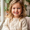 blonde_hair, blue_eyes, bookshelf, casual_clothing, child, cozy, cute, girl, happy, home, indoor, knitwear, natural_light, plants, portrait, sitting, smiling, sweater, window, young
