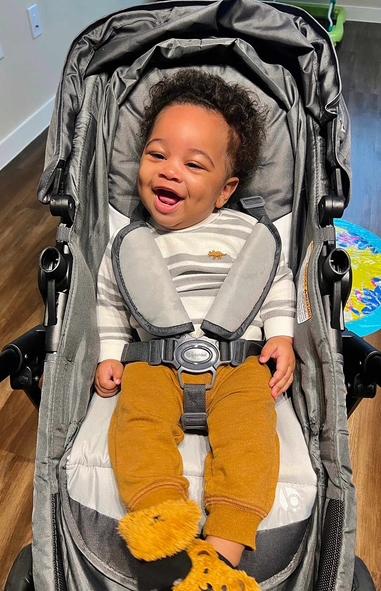 Landon joined the competition — help win amazing prizes! baby, baby_clothing, child, curly_hair, cute, fuzzy_socks, happy, home, indoor, infant, mustard_pants, person, playmat, portrait, seatbelt, sitting, smiling, striped_shirt, stroller, wood_floor