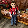 Aaron a rejoint le concours — aidez-le/la à gagner de superbes lots ! baby_bottle, blanket, boy, brown_couch, child, christmas_tree, clothing, decorations, face, furniture, holiday, home, indoor, jeans, person, red_shirt, standing, suspenders, toy, wood_floor