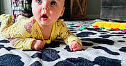 Bryce is registered to the contest to win money with this photo: baby, black_and_white_pattern, bright_eyes, cabinet, carpet, child, couch, curious_expression, face, floor, furniture, hands, head, indoor, infant, living_room, lying_down, person, toys, yellow_pajamas