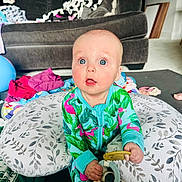 Bryce joined the competition — help win amazing prizes! baby, child, indoors, living_room, couch, pillow, toy, onesie, flamingo_pattern, curious_expression, floor_mat, blanket, clothing, person, home, playtime, sitting, blue_eyes, infant, cute