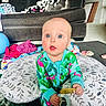 baby, child, indoors, living_room, couch, pillow, toy, onesie, flamingo_pattern, curious_expression, floor_mat, blanket, clothing, person, home, playtime, sitting, blue_eyes, infant, cute