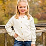 child, girl, smile, blonde_hair, white_sweater, blue_jeans, outdoor, autumn, wooden_railing, nature, portrait, happy, casual_clothing, posing, young, person, fashion, fall_foliage, daylight, cute
