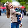 child, baby, girl, smiling, holding, outdoor, street, denim, overalls, bow, hair_accessory, casual_clothing, happy, sibling, family, portrait, shoes, greenery, blurred_background, people