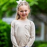 child, girl, smiling, outdoor, nature, greenery, long_sleeve_shirt, heart_detail, blue_jeans, hair_bow, portrait, happy, standing, watch, casual_clothing, daylight, cute, fashion, person, young