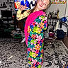 child, girl, floral_dress, pink_scarf, living_room, carpet, television, furniture, bottle, chair, toy, shelf, plant, backpack, rug, smile, pose, home_interior, person, decor
