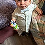 toddler, child, boy, indoor, shirt, bow_tie, juice_bottle, toy, floor, quilt, couch, carpet, curious, standing, furniture, face, clothing, hand, person, expression