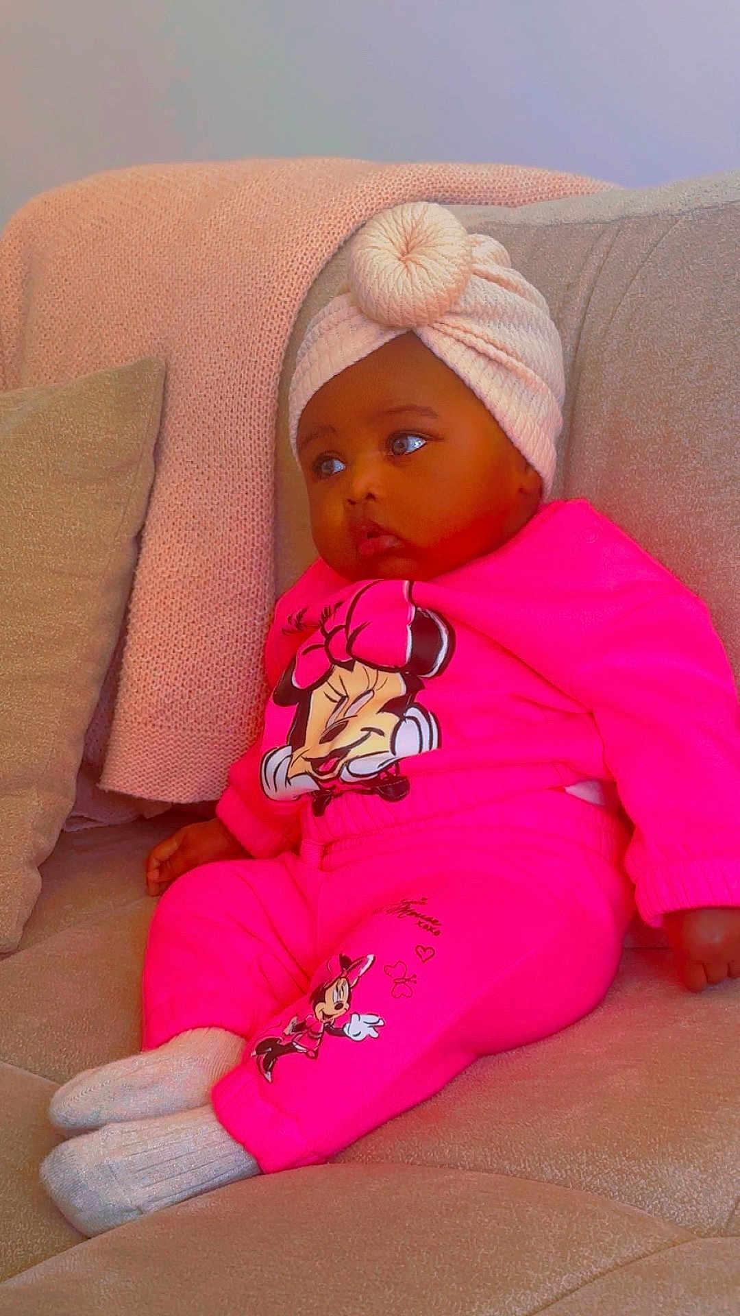 Miya a rejoint le concours — aidez-le/la à gagner de superbes lots ! baby, child, infant, pink_clothing, turban, couch, blanket, pillow, socks, minnie_mouse, cartoon_character, indoor, seated, cute, soft_texture, cozy, portrait, casual, relaxed, looking_away