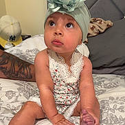 Olive is registered to the contest to win money with this photo: baby, bed, blanket, child, cute, expression, flower, foot, hand, headband, indoors, infant, looking_up, person, pillow, romper, sitting, skin, tattoo, young