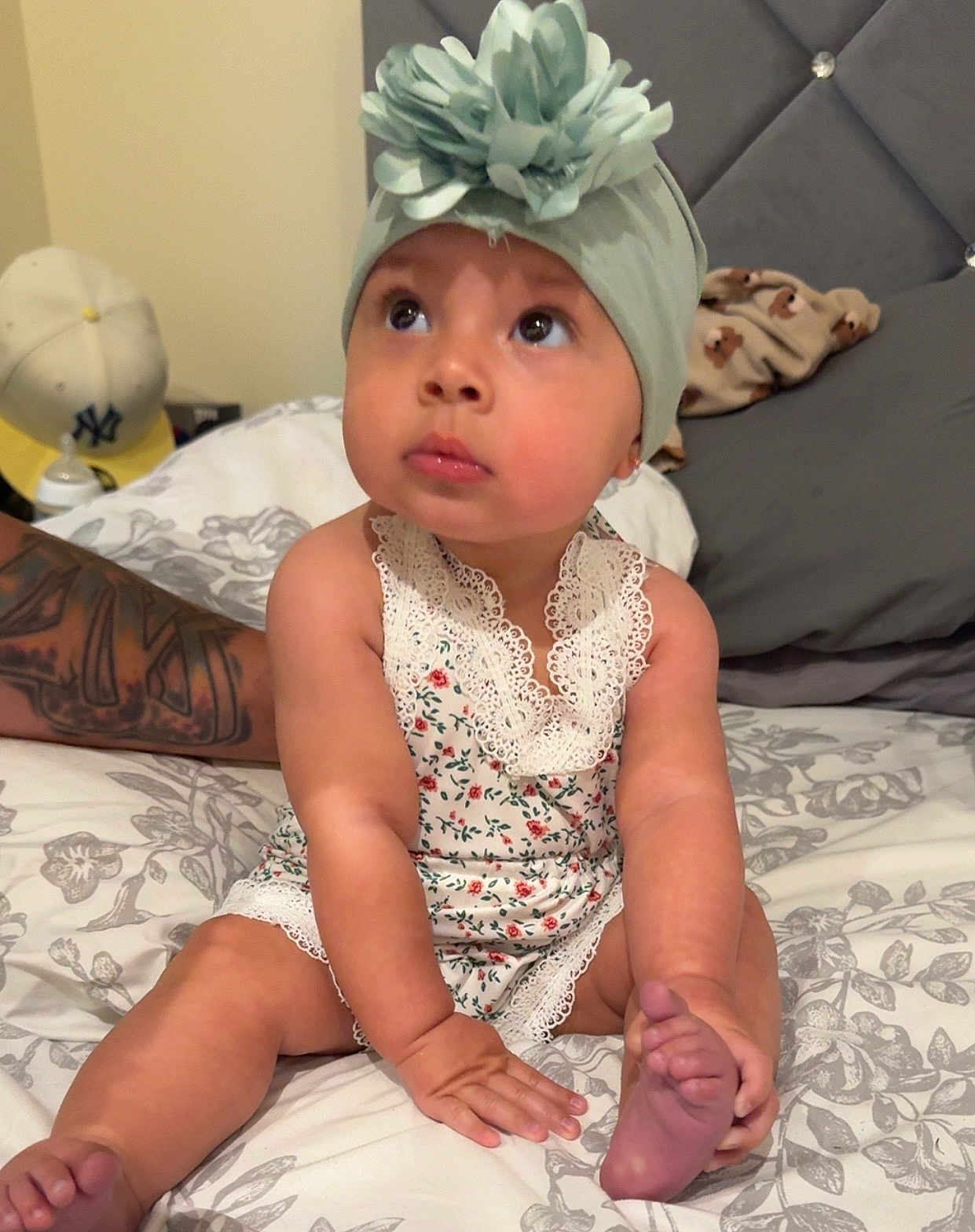 Olive is registered to the contest to win money with this photo: baby, bed, blanket, child, cute, expression, flower, foot, hand, headband, indoors, infant, looking_up, person, pillow, romper, sitting, skin, tattoo, young