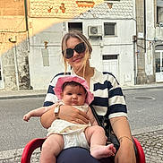 Ambre a rejoint le concours — aidez-le/la à gagner de superbes lots ! woman, baby, child, sunglasses, hat, outdoor, building, street, table, chair, smile, person, sunlight, casual_clothing, accessories, watch, bracelet, pavement, daytime, portrait