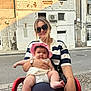 woman, baby, child, sunglasses, hat, outdoor, building, street, table, chair, smile, person, sunlight, casual_clothing, accessories, watch, bracelet, pavement, daytime, portrait