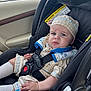 baby, child, car_seat, car_interior, seat_belt, clothing, headwear, expression, face, person, sitting, safety, vehicle, window, road, trees, daylight, cute, infant, transport