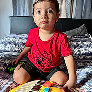 Mcallister is registered to the contest to win money with this photo: bed, black_shorts, casual_clothing, child, curly_hair, cute, daylight, expression, furniture, hands, home, indoor, patterned_bedspread, playing, portrait, red_shirt, sitting, toddler, toy_keyboard, young_child
