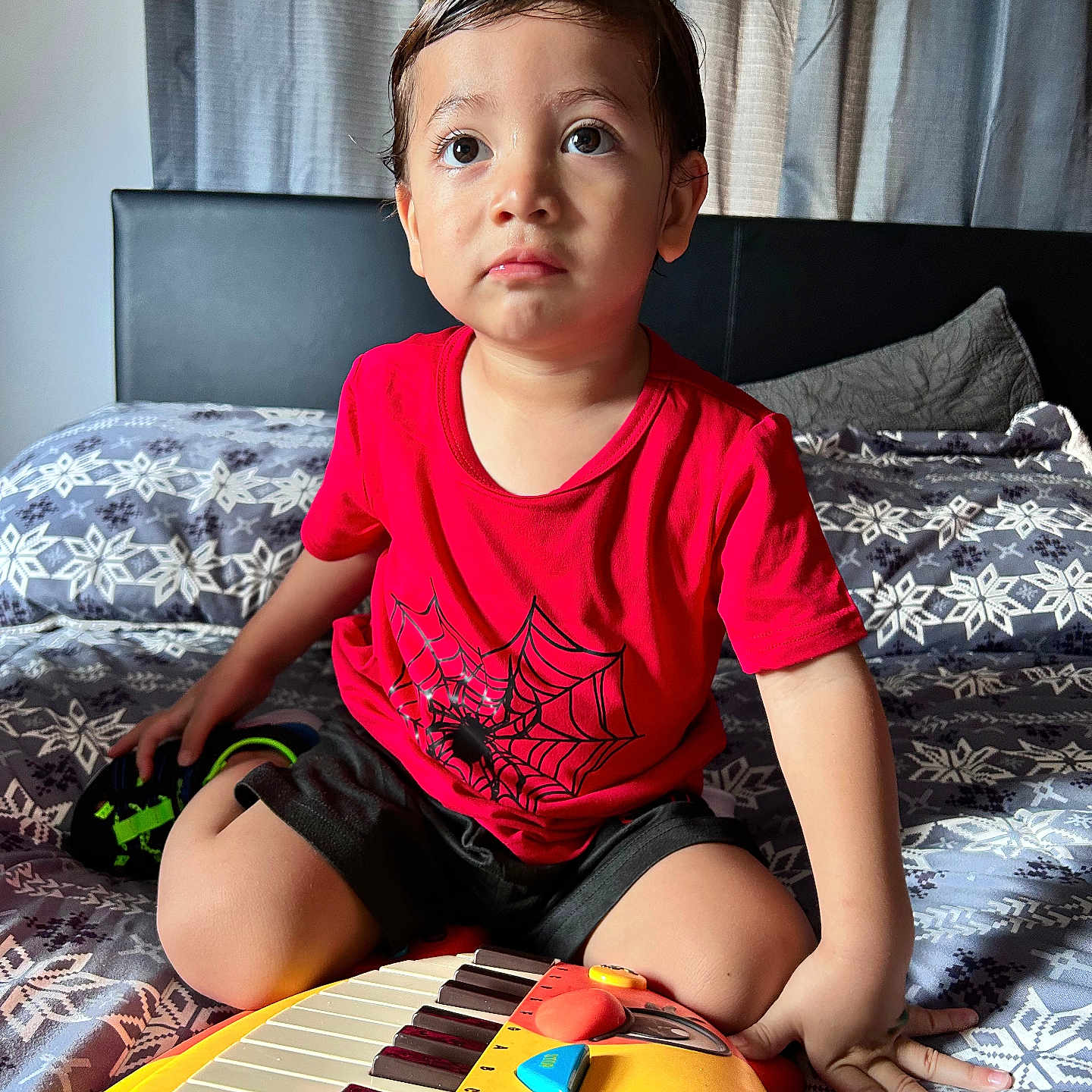 Mcallister is registered to the contest to win money with this photo: bed, black_shorts, casual_clothing, child, curly_hair, cute, daylight, expression, furniture, hands, home, indoor, patterned_bedspread, playing, portrait, red_shirt, sitting, toddler, toy_keyboard, young_child