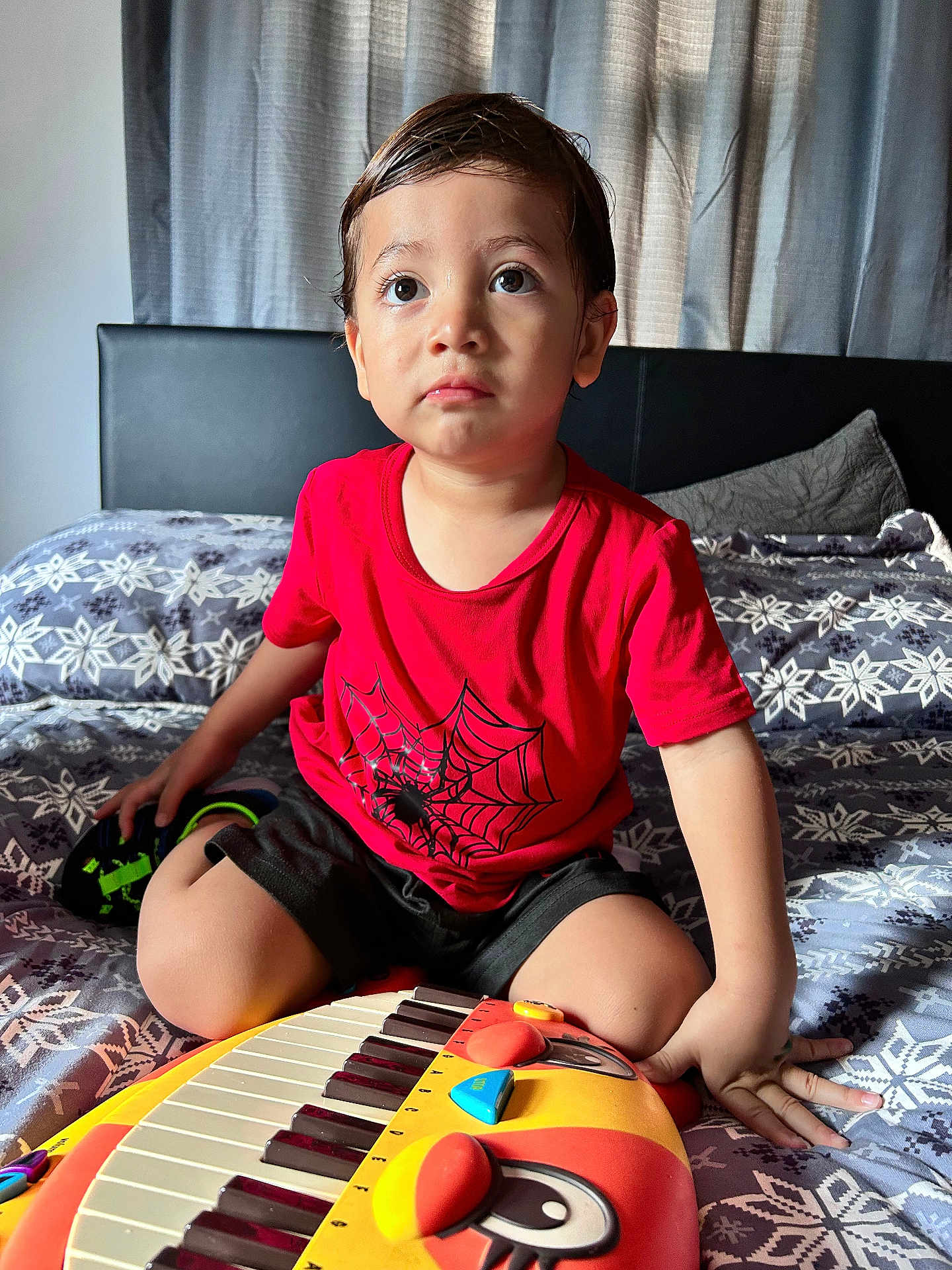 Mcallister is registered to the contest to win money with this photo: bed, black_shorts, casual_clothing, child, curly_hair, cute, daylight, expression, furniture, hands, home, indoor, patterned_bedspread, playing, portrait, red_shirt, sitting, toddler, toy_keyboard, young_child