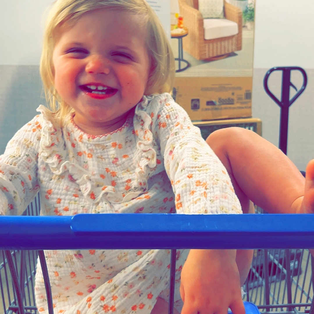 Paisleigh is registered to the contest to win money with this photo: toddler, child, smiling, shopping_cart, indoor, box, lounge_chair, blonde_hair, dress, happy, playful, person, retail, floor, hand, foot, metal, wire, cute, young