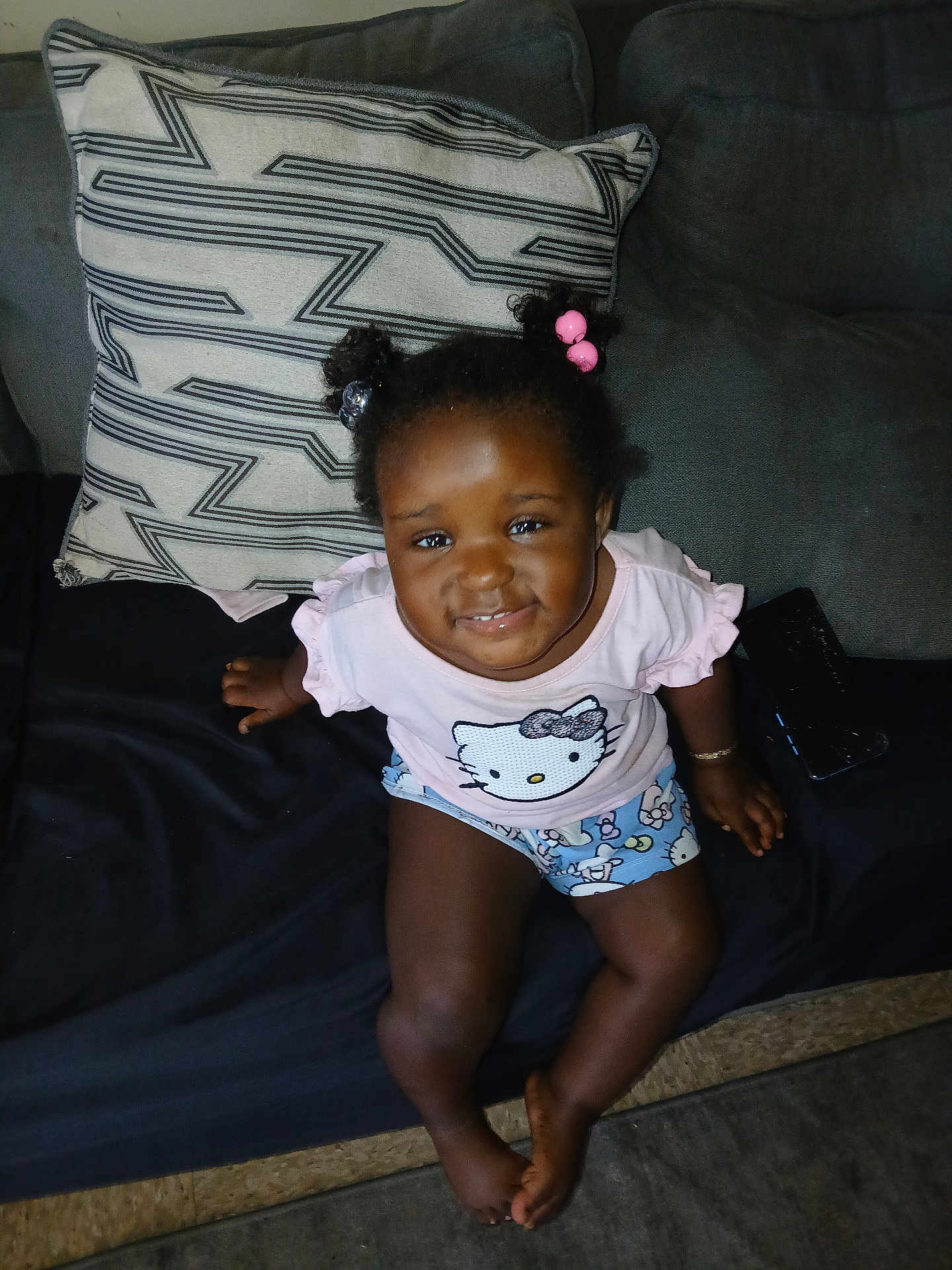 Shameria is registered to the contest to win money with this photo: child, toddler, smiling, sitting, couch, pillow, hair_beads, pink_shirt, hello_kitty, shorts, indoor, cute, happy, person, young_child, curly_hair, barefoot, living_room, portrait, casual_clothing
