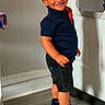 black_shorts, boy, casual_clothing, child, cute, happy, home, indoor, mirror, portrait, posing, red_cap, reflection, shorts, smiling, sneakers, socks, standing, wall_art, wooden_floor