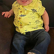 Hunter is registered to the contest to win money with this photo: baby, child, smiling, yellow_shirt, dinosaurs, jeans, white_sneakers, sitting, couch, indoor, happy, cute, portrait, infant, clothing, footwear, person, young_child, casual, relaxed