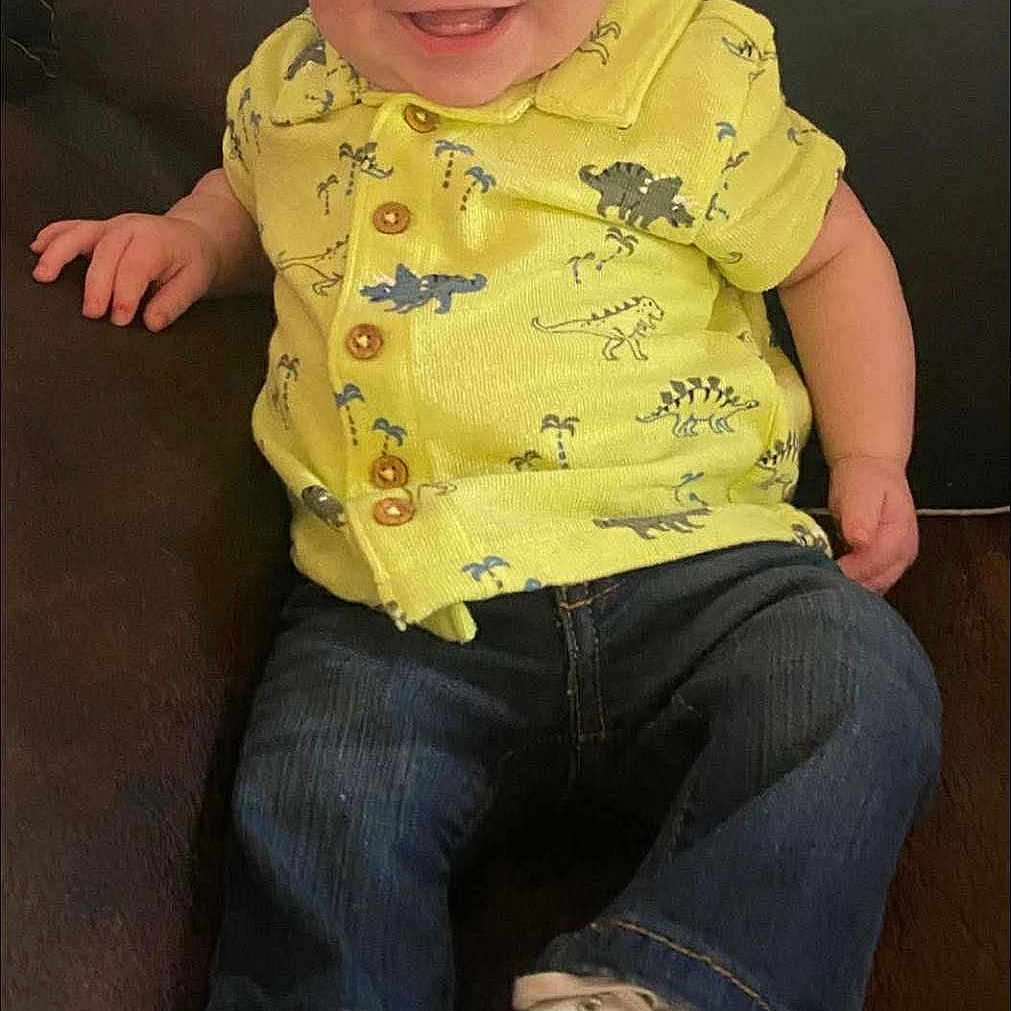 Hunter is registered to the contest to win money with this photo: baby, casual, child, clothing, couch, cute, dinosaurs, footwear, happy, indoor, infant, jeans, person, portrait, relaxed, sitting, smiling, white_sneakers, yellow_shirt, young_child