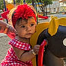 Alanii is registered to the contest to win money with this photo: baby, child, toddler, playground, carousel, ride_on_toy, bull_ride, flower_headband, red_dress, gingham, smiling, portrait, outdoor, park, trees, sunlight, play_equipment, seated, summer, colorful