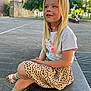 child, girl, blonde_hair, tshirt, skirt, leopard_print, sandals, sitting, outdoor, bench, stone, smiling, daylight, trees, building, casual, happy, young, portrait, park