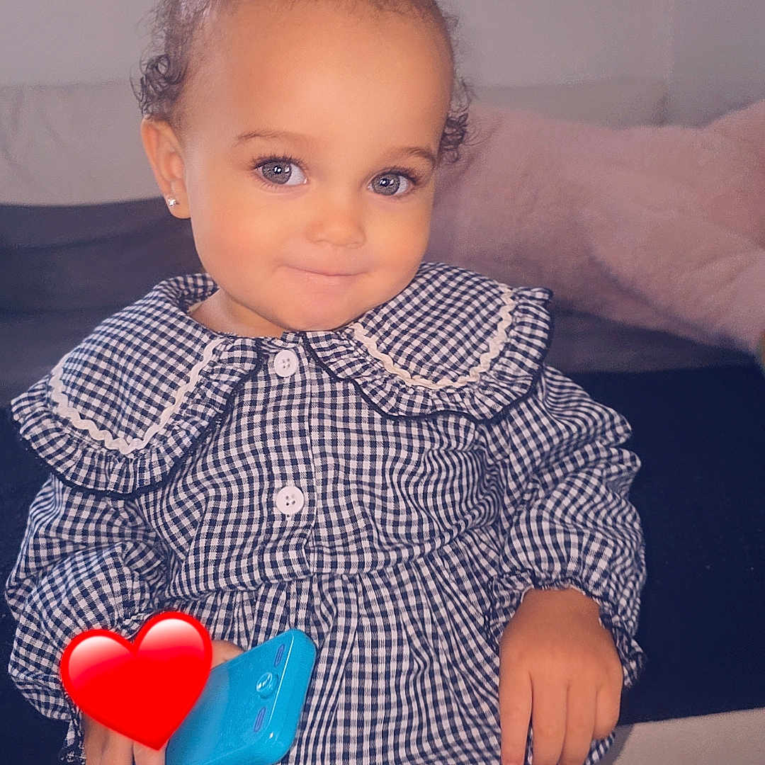 Ava a rejoint le concours — aidez-le/la à gagner de superbes lots ! blue_toy_phone, checkered_clothing, child, couch, cozy, curly_hair, cute, earrings, furniture, hair_buns, happy, home, indoor, pajamas, person, playing, portrait, smiling, standing, toddler