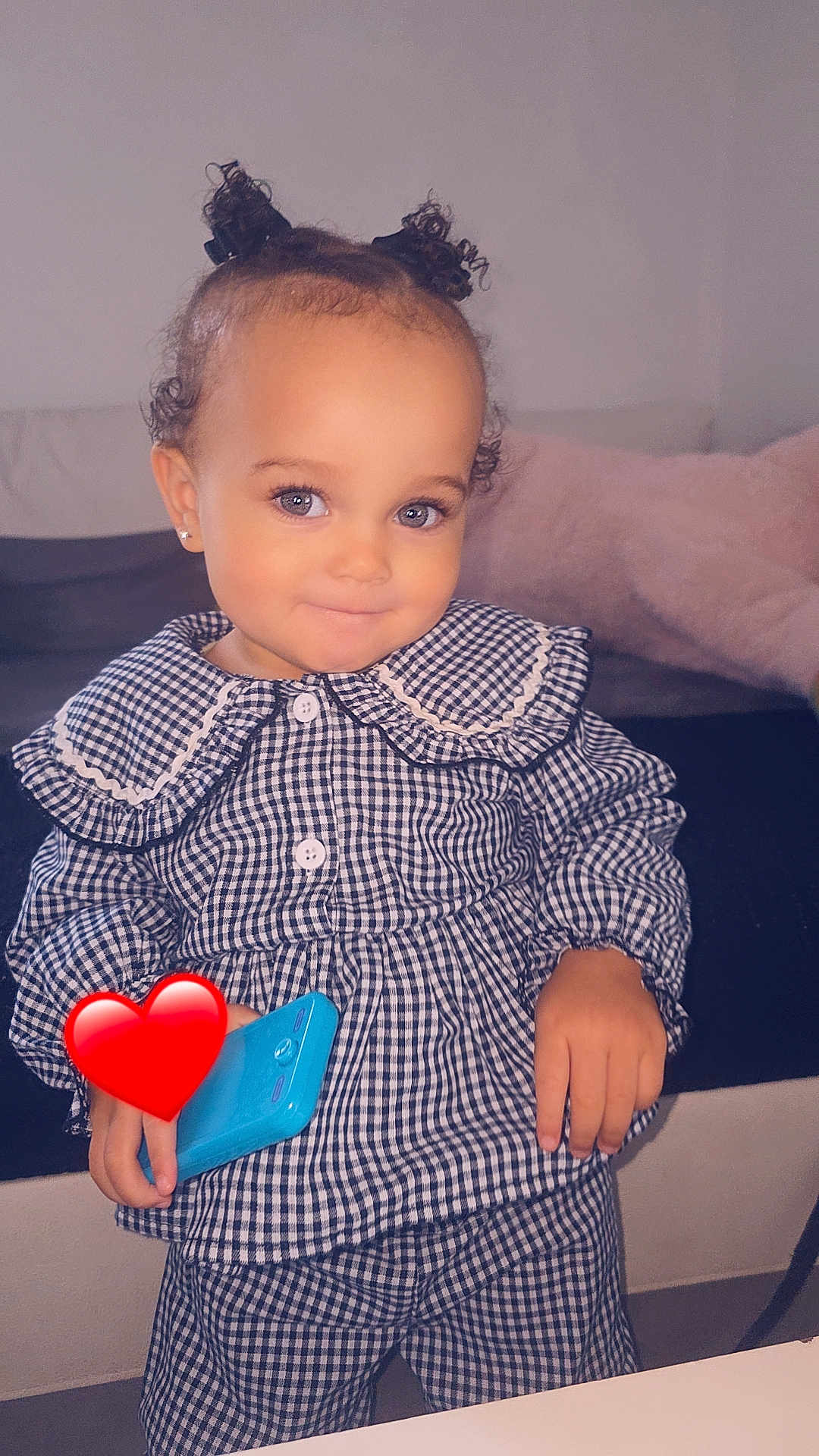 Ava a rejoint le concours — aidez-le/la à gagner de superbes lots ! toddler, child, smiling, curly_hair, indoor, checkered_clothing, blue_toy_phone, cute, portrait, person, furniture, couch, pajamas, hair_buns, earrings, happy, standing, playing, home, cozy