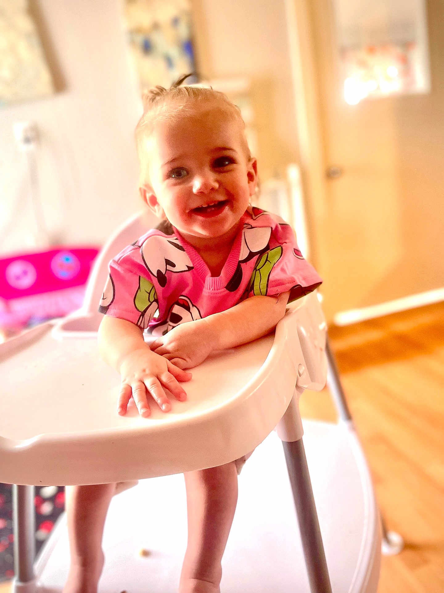 Mia joined the competition — help win amazing prizes! toddler, child, high_chair, smiling, pink_shirt, indoor, person, happy, face, small_hands, legs, wooden_floor, furniture, home, bright, cute, baby, cartoon_print, hair_tie, playful
