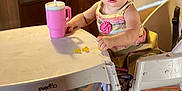 Mia is registered to the contest to win money with this photo: toddler, child, high_chair, sippy_cup, gummy_bears, table, play_mat, carpet, wooden_furniture, indoor, person, face, blue_eyes, clothing, plastic, cup, floor, hand, foot, curious