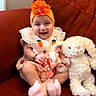 baby, smile, orange_headband, flower, carrot_dress, stuffed_bunny, plush_toy, red_couch, happy, child, indoors, cute, infant, soft_toy, sitting, portrait, headwear, toy, cozy, playful
