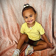Nevaeh is registered to the contest to win money with this photo: child, toddler, smiling, portrait, sitting, yellow_shirt, hair_bow, sneakers, blanket, indoors, happy, face, person, fashion, curly_hair, brown_skin, legs, casual_clothing, studio_portrait, fabric_background