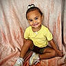 child, toddler, smiling, portrait, sitting, yellow_shirt, hair_bow, sneakers, blanket, indoors, happy, face, person, fashion, curly_hair, brown_skin, legs, casual_clothing, studio_portrait, fabric_background