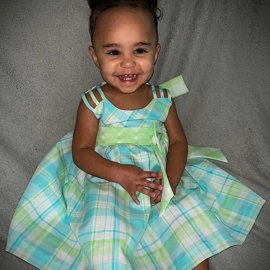 Nevaeh is registered to the contest to win money with this photo: child, toddler, girl, smiling, portrait, sitting, dress, plaid, colorful, indoors, face, hair_bows, happy, fashion, baby, feet, gray_background, fabric, photography, cute