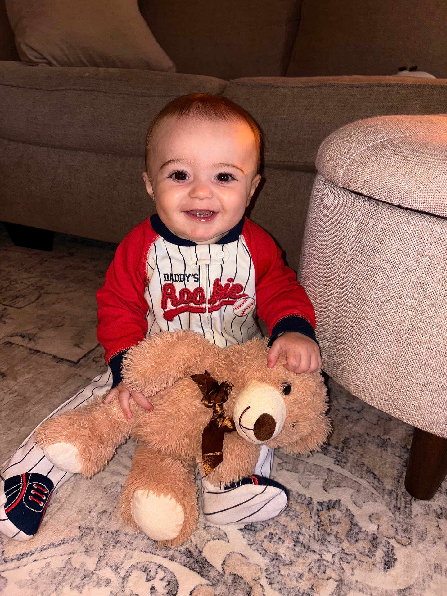 Grayson joined the competition — help win amazing prizes! baby, child, teddy_bear, plush_toy, smiling, sitting, indoor, carpet, couch, ottoman, clothing, onesie, footwear, happy, young_child, person, home, cute, soft_toy, floor