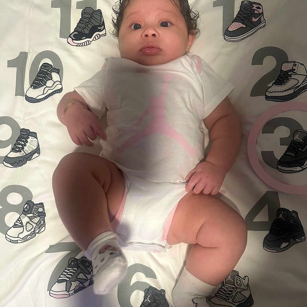 Dayanna joined the competition — help win amazing prizes! baby, infant, child, onesie, socks, curly_hair, blanket, numbers, sneakers, footwear, laying_down, cute, indoor, portrait, soft_lighting, shadow, playful, clothing, person, young_child