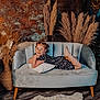 child, girl, smiling, sofa, pillow, dress, braided_hair, pampas_grass, vase, wooden_floor, rug, indoor, portrait, happy, resting, furniture, home_decor, cozy, freckles, plush